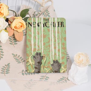 Könnte beinhalten: Ein rechteckiger Dekorationsartikel mit dem Text "THE NEW YORKER" oben. Das Design zeigt einen grünen Hintergrund mit vertikalen weißen Streifen und Illustrationen von Katzenpfoten, die nach Spaghetti greifen. Der Artikel ist auf einem Stoff mit floralem Muster platziert.