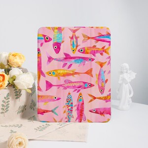 Könnte beinhalten: Eine Tablet-Hülle mit einem Aquarelldesign von bunten Fischen in Rosa-, Orange-, Gelb- und Blautönen auf einem rosa Hintergrund. Die Fische sind in verschiedenen Richtungen angeordnet und erzeugen ein lebendiges und verspieltes Muster.