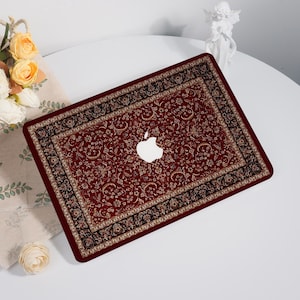 May include: A laptop skin featuring a deep red background with an intricate black and gold floral pattern reminiscent of a traditional rug design. The skin has a central cutout revealing the white Apple logo on the laptop lid surface.