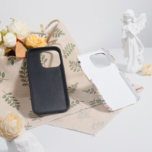 May include: Two phone cases, one black and one white, are displayed. The black case is solid, while the white case has a floral design. They are placed on a beige cloth with green leaf patterns, near a basket of flowers and a white angel statue.