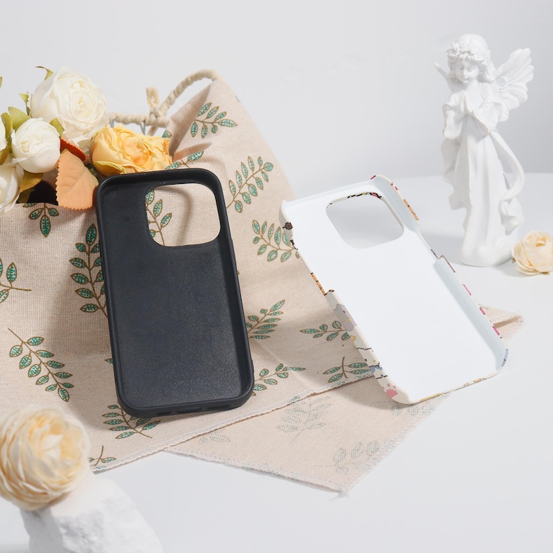 May include: Two phone cases, one black and one white, are displayed. The black case is solid, while the white case has a floral design. They are placed on a beige cloth with green leaf patterns, near a basket of flowers and a white angel statue.