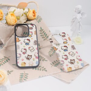 May include: Two phone cases with a white background and a repeating pattern of Hello Kitty characters in various poses. One case has a black border. The cases are on a beige cloth with green leaf designs.