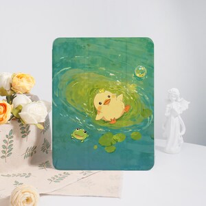 May include: A tablet case featuring a whimsical illustration of a yellow duckling floating in a pond with a green frog and lily pads. The background is a watercolor-style depiction of water with bubbles.