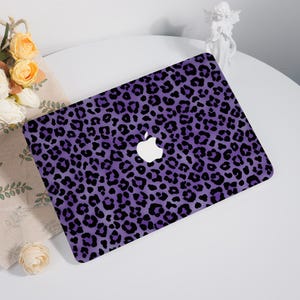 May include: A laptop with a purple leopard print design. The laptop has a white Apple logo in the center. The background is a white table with flowers and a small statue.