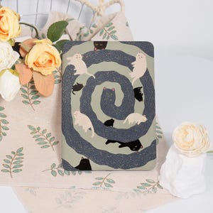 May include: A rectangular, cat-themed tablet case featuring a whimsical design. The case has a light green background with a dark blue, swirling pattern. White and black cats are scattered throughout the design. The case is surrounded by a floral arrangement and a decorative cloth.