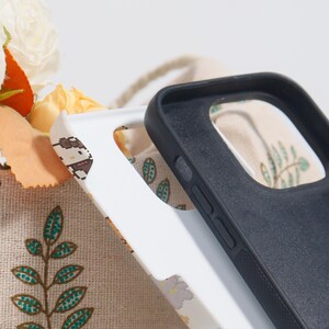 May include: Close-up of two phone cases. One is white with a pixelated design, the other is black. The cases are made of a smooth material and have cutouts for the phone's buttons and camera. The background includes a floral pattern.