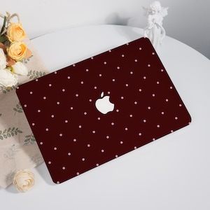 Könnte beinhalten: Eine weinrote Laptop-Hülle mit einem Muster aus kleinen, gleichmäßig verteilten rosa Punkten. Das Apple-Logo ist weiß. Die Hülle liegt auf einer weißen Oberfläche, mit Blumen und einer kleinen Statue im Hintergrund.