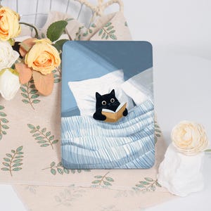 May include: A rectangular art print depicts a black cat reading a book in bed. The cat is under a blue and white striped blanket, with white pillows. The background is a light blue, with a hint of sunlight.