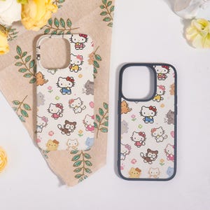 May include: Two phone cases with a repeating pixelated Hello Kitty pattern in pink, blue, yellow, and brown on a cream background. One case has a white border, and the other has a dark blue border. The cases are on a beige cloth with green leaf patterns.