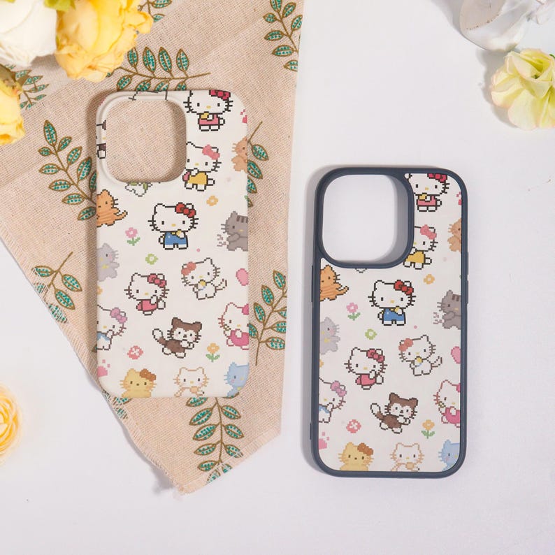 May include: Two phone cases with a repeating pixelated Hello Kitty pattern in pink, blue, yellow, and brown on a cream background. One case has a white border, and the other has a dark blue border. The cases are on a beige cloth with green leaf patterns.