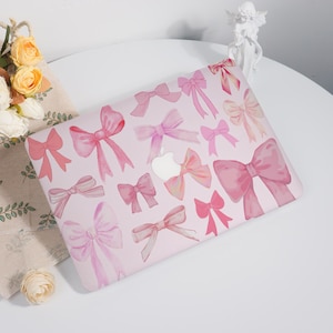 May include: A pink laptop case decorated with watercolour bows in different shades of pink. The bows are spread across the case, creating a whimsical design. The Apple logo is visible in the centre. The case is on a white surface.