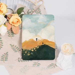 May include: A rectangular e-reader case featuring a printed illustration of a person sitting on a dark green hill overlooking golden mountains under a cloudy sky with a small black cross centered in the distance against a bright light source.
