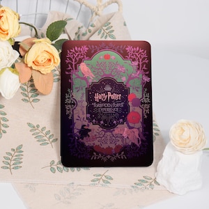 May include: A rectangular, dark-colored item with the words "Harry Potter Forbidden Forest Experience" in gold lettering. The design features a forest scene with an owl, trees, and other magical elements. The item is placed on a floral patterned cloth.