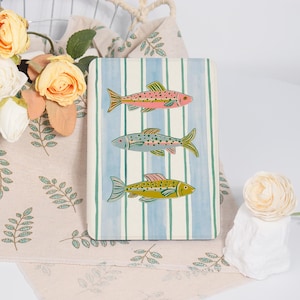 May include: A rectangular decorative board with a blue and white striped background. Three colorful fish are painted on the board: pink, gray, and yellow. The fish have detailed fins and scales. The board is placed on a floral patterned fabric.