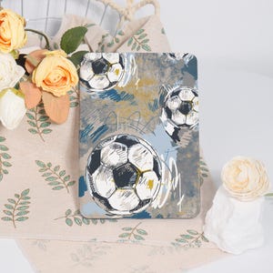 May include: A rectangular tablet case featuring a soccer ball design. The case has a gray background with blue and yellow brushstrokes and multiple soccer ball illustrations. The soccer balls are black and white with yellow accents.