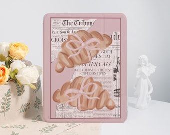 Könnte beinhalten: Eine rosa Tablet-Hülle mit einem Croissant-Design und einer rosa Schleife. Der Hintergrund zeigt einen Zeitungsdruck mit dem Text "The Tribun" und "Corner Cafe". Die Hülle befindet sich vor einem weißen Hintergrund mit Blumen und einer Statue.