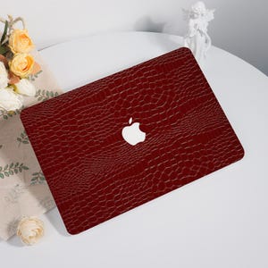 May include: A burgundy laptop cover with a textured, crocodile-skin pattern. The cover features the Apple logo in white. The laptop is placed on a white surface, with flowers and a small statue in the background.