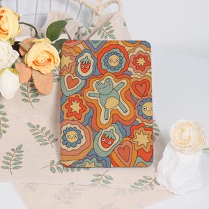 May include: A rectangular, colorful case featuring a retro design with a light blue frog, flowers, hearts, and strawberries. The case has a groovy, psychedelic style with orange, blue, and red hues.