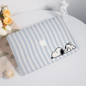 May include: A light blue and white striped laptop cover featuring a white Apple logo and a black and white Snoopy graphic. The Snoopy graphic depicts Snoopy sleeping. The cover is designed to fit a laptop.