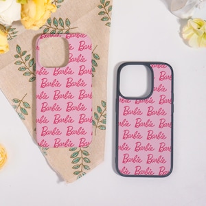 May include: Two pink phone cases with a repeating "Barbie" script in red. One case is on a patterned beige cloth with green leaf designs. The other case has a black border.