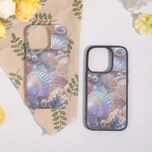 May include: Two phone cases with a seashell design. The cases feature a clear background with iridescent seashells, pearls, and coral. One case has a clear border, and the other has a black border. The cases are on a beige cloth with green leaf patterns.