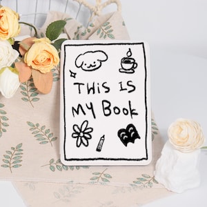 Peut inclure: Une couverture de livre blanche avec des gribouillis noirs et le texte "THIS IS MY Book". Les gribouillis comprennent un chien, une tasse de café, une fleur, un crayon et un cœur. Le livre est entouré de fleurs.
