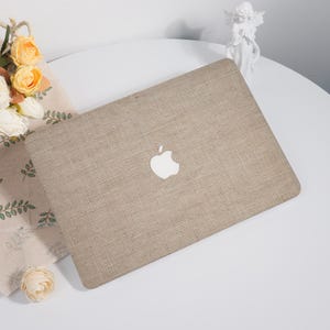 May include: A beige laptop cover with a textured burlap design and a white Apple logo. The cover is on a white surface, with a floral arrangement and a small statue in the background.
