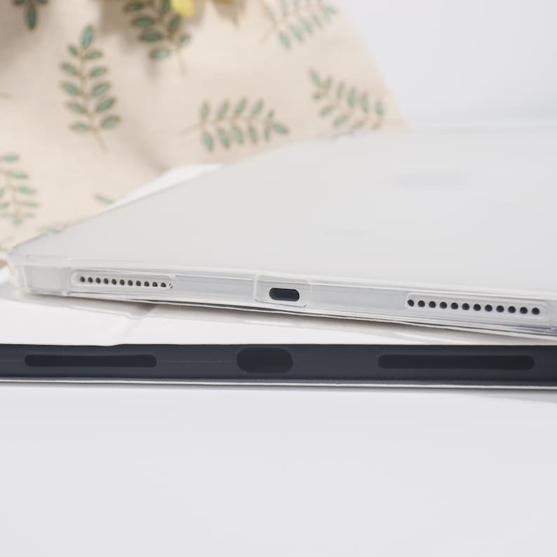 K&ouml;nnte beinhalten: Eine durchsichtige Schutzh&uuml;lle f&uuml;r ein Tablet mit pr&auml;zisen Aussparungen f&uuml;r Lautsprecher und einen zentralen Ladeanschluss. Die H&uuml;lle liegt auf einer schwarzen Tablet-Abdeckung und zeigt das schlanke Profil sowie das funktionale Design.