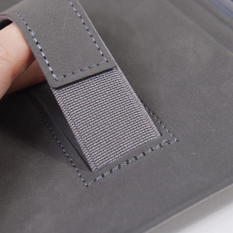 May include: Close-up of a grey notebook with a fabric handle. The handle is attached to the notebook with grey stitching. The notebook is a solid grey colour.