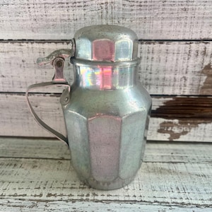 May include: A silver metal creamer with a octagonal shape and a hinged lid. The creamer has a handle and a spout.