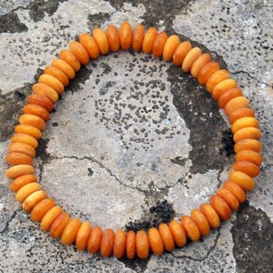 Op de afbeelding: Een cirkelvormige armband bestaande uit kleine, afgeronde oranje kralen. De kralen zijn aan elkaar geregen en vormen een doorlopende lus. De armband ligt op een getextureerd, grijs stenen oppervlak.