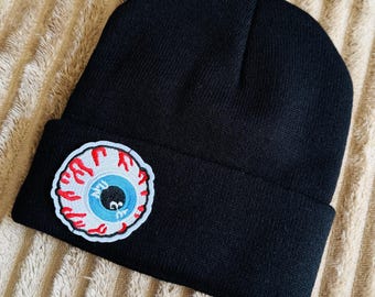 May include: A black knit beanie featuring a circular embroidered patch on the cuff. The patch depicts a realistic human eyeball with a blue iris, a black pupil, and prominent red blood vessels radiating outward against a white background.