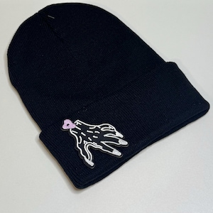 May include: Black knit beanie with a hand graphic. The hand graphic is black with white outlines and silver fingernails. The graphic has a pink heart on the wrist.