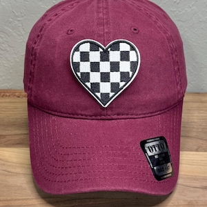 May include: A maroon baseball cap featuring a heart-shaped patch with a black and white checkered pattern. The cap has a curved brim and a black sticker on the lower right side with the text OTTO, RELAXED FIT, and DAD HAT printed in white.