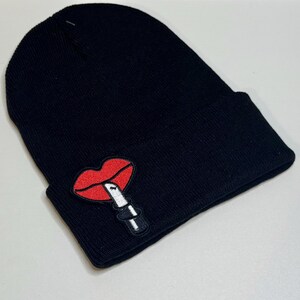May include: Black knit beanie featuring a red lips and lipstick patch. The patch is embroidered with a detailed design. The beanie is a classic style, suitable for cold weather and casual wear.