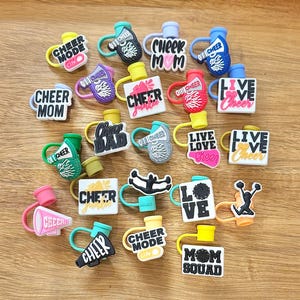 May include: A collection of 18 colorful cheerleading themed bottle stoppers. The stoppers feature various cheerleading slogans, including "Cheer Mode", "Cheer Mom", "Live Love Cheer", and "Cheer Squad".