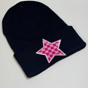 May include: Black beanie hat with a pink star patch. The star features a checkered pattern in shades of pink and a white border. The hat is made of a soft, knitted material.