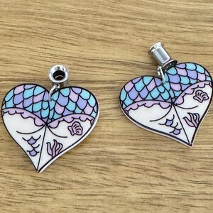 May include: A pair of heart-shaped earrings with a mermaid scale design in shades of blue and purple. The earrings have a white base with black outlines and silver-colored metal findings. The design includes seashell and seaweed motifs.