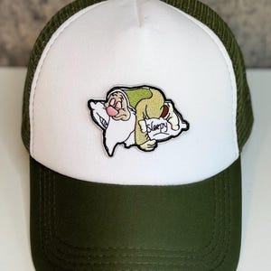 May include: A green and white trucker hat featuring an embroidered patch of the Disney character Sleepy from Snow White. The patch shows Sleepy sleeping on a pillow with the word "Sleepy" on a sign. The hat has a green mesh back and a white front.
