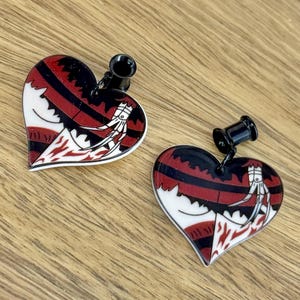 May include: A pair of heart-shaped earrings with a black, red, and white design. The earrings feature a black plug and a detailed illustration. The design includes black stripes, white accents, and a red and white graphic.