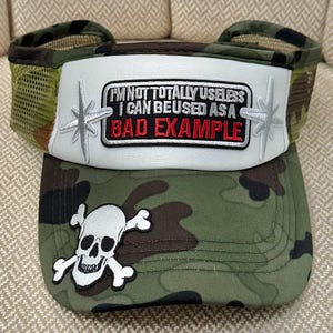 May include: A camouflage trucker hat with a white front panel featuring the text "I'M NOT TOTALLY USELESS I CAN BE USED AS A BAD EXAMPLE." The hat has a skull and crossbones patch and a mesh back.