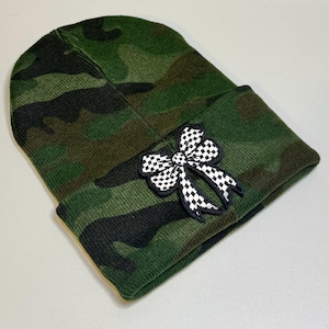 May include: A camouflage-patterned beanie hat with a black and white checkered bow applique. The hat features a folded brim and is primarily green, brown, and black. The bow is a contrasting detail.