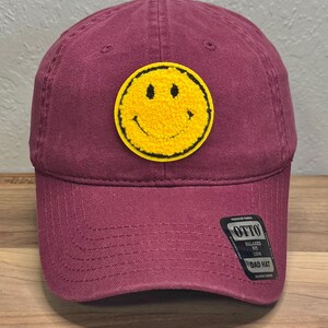 May include: A maroon baseball cap featuring a circular yellow tufted smiley face patch on the front. A black sticker on the brim displays the text OTTO, RELAXED FIT, OSFM, and DAD HAT in white lettering, with additional small text.