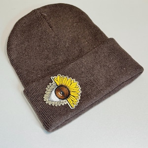 May include: A brown knit beanie with a folded brim. The beanie features a patch of a sunflower with an eye in the center. The sunflower has yellow petals and a brown center. The eye has a white sclera and a brown iris.