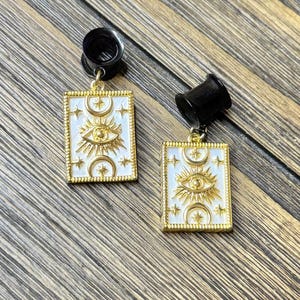 May include: A pair of black ear plugs with dangling rectangular gold-toned charms. Each charm features a white enamel background with a central gold eye motif, surrounded by crescent moon and star symbols, all framed by a textured gold border.