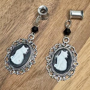 May include: A pair of silver-toned cat cameo earrings. Each earring features a white cat silhouette on a black background within an ornate oval frame. A black bead and chain connect the frame to the ear plug.