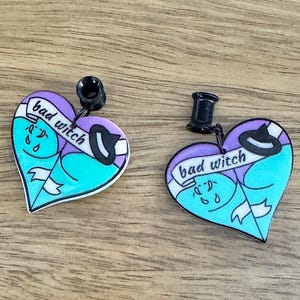 May include: A pair of heart-shaped earrings with a witch theme. Each earring features a purple heart with a teal interior, a witch hat, and the words "bad witch" on a banner. The earrings have black posts.