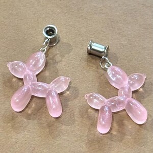 May include: Two pink balloon dog earrings. Each earring features a clear, pink, dog-shaped charm suspended from a silver-colored post. The earrings are displayed on a neutral-toned surface.