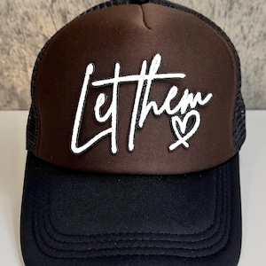 May include: A brown and black trucker hat with the phrase "Let them" in white script, accompanied by a heart symbol. The hat features a mesh back and a curved brim.