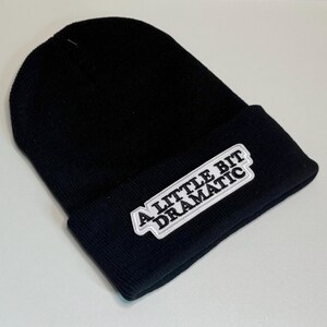 May include: Black beanie hat with a folded brim. A white and gray patch on the front reads "A LITTLE BIT DRAMATIC". The hat is made of a soft, knitted material.